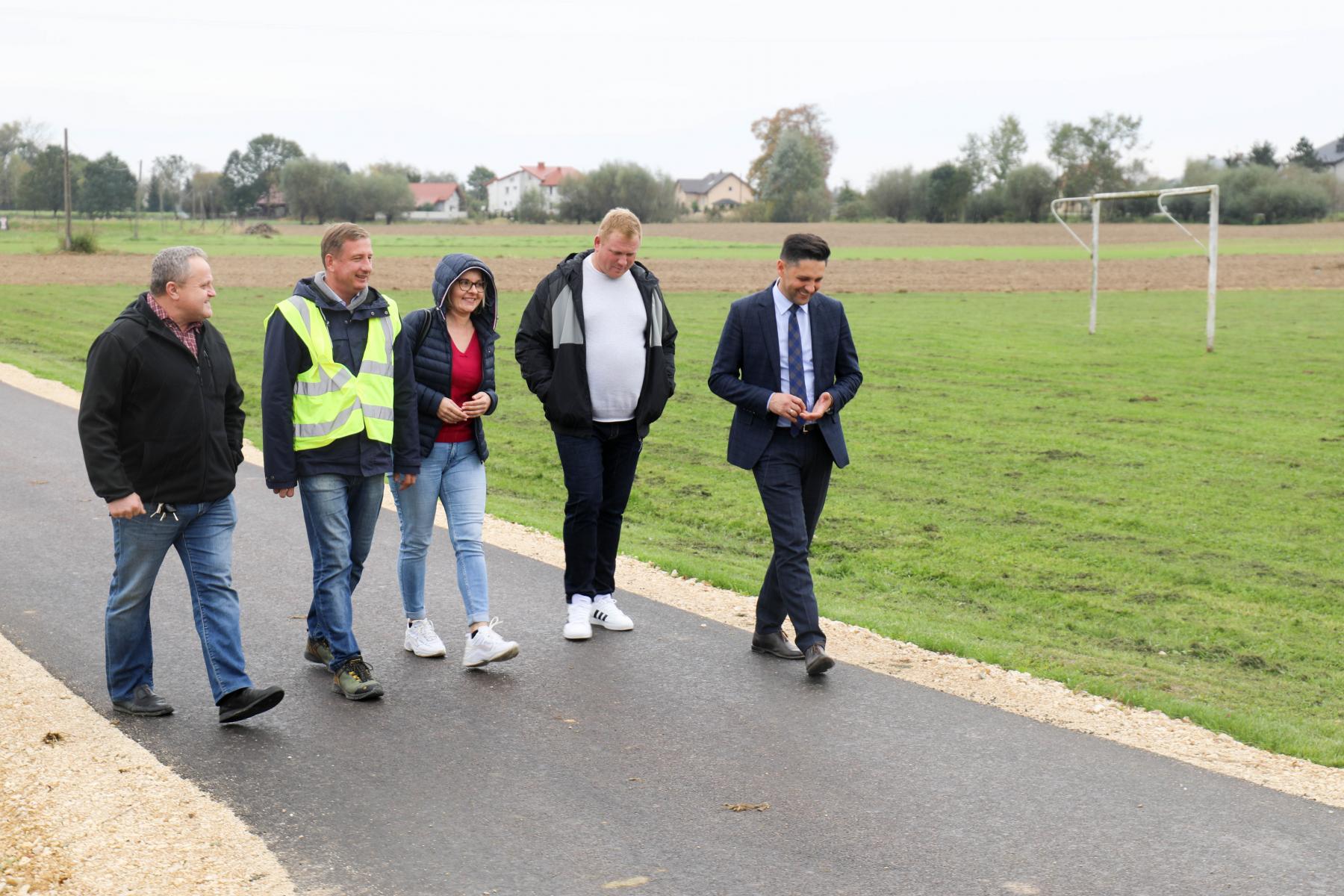 Na zdjęciu znajdują się osoby, droga i tablica informacyjna.